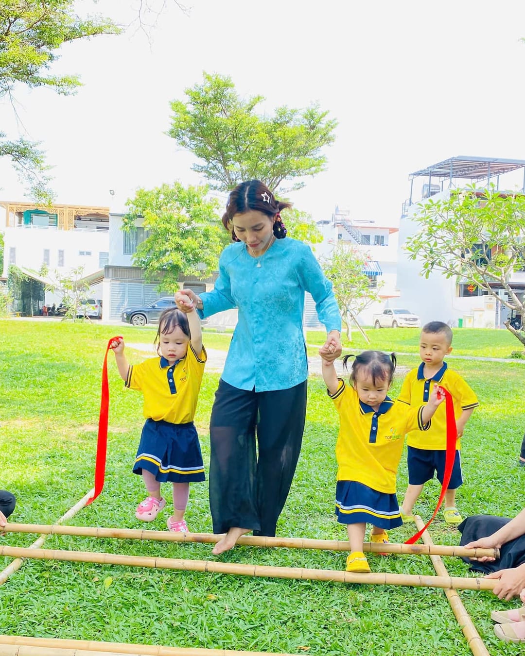 Cô giáo trong tà áo bà ba nắm tay hai bé bước qua trò chơi múa sạp trên bãi cỏ xanh
